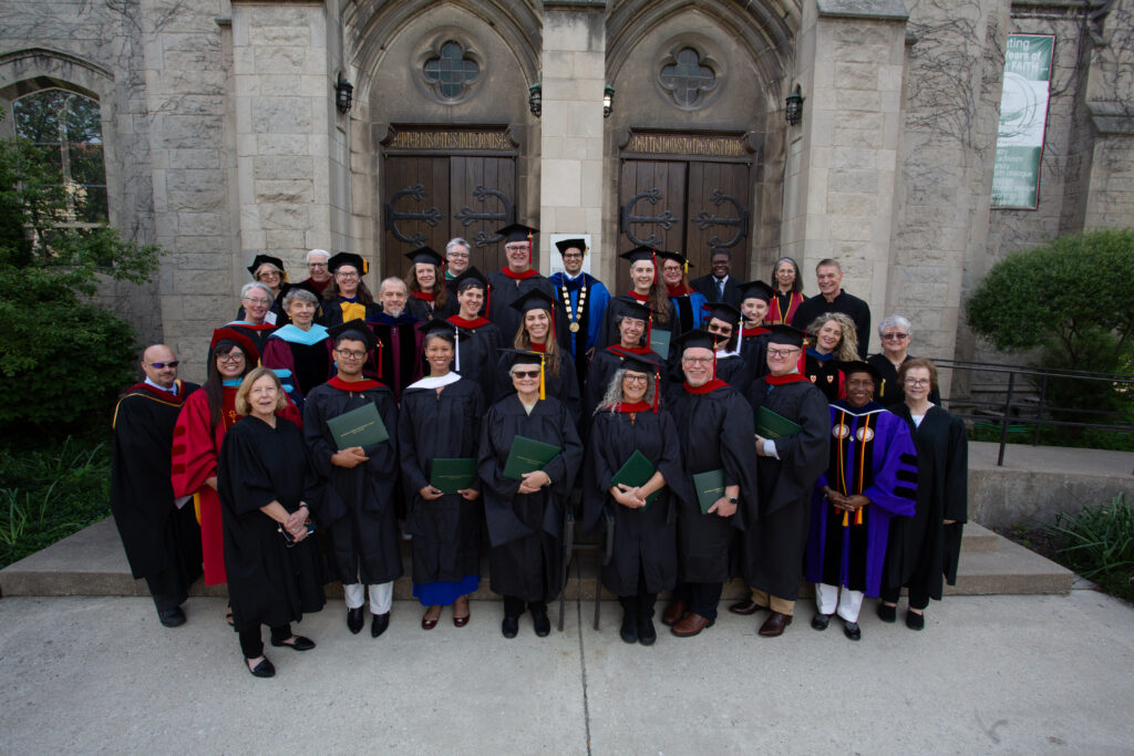 Group portrait of approximately 40 faculty and staff members wearing academic regalia (black robes, colorful hoods, and mortarboards) standing in formal rows in front of a Gothic stone building with two large arched doorways, ornate doors, and circular windows. Many are holding green diplomas or documents. The setting appears to be a university or academic institution.